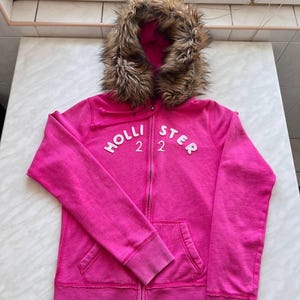 May include: A bright pink zip-up hoodie with a faux fur-lined hood. The hoodie features the brand name "HOLLISTER" in white block letters across the chest, with the number "22" below. It has a front pocket and ribbed cuffs.