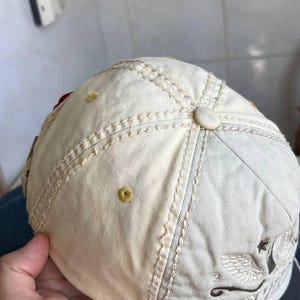 Op de afbeelding: Cr&egrave;mekleurige baseballpet met een gebogen klep. De pet heeft decoratieve stiksels, een knoop aan de bovenkant en een geborduurd ontwerp aan de zijkant. De pet wordt door een hand vastgehouden.