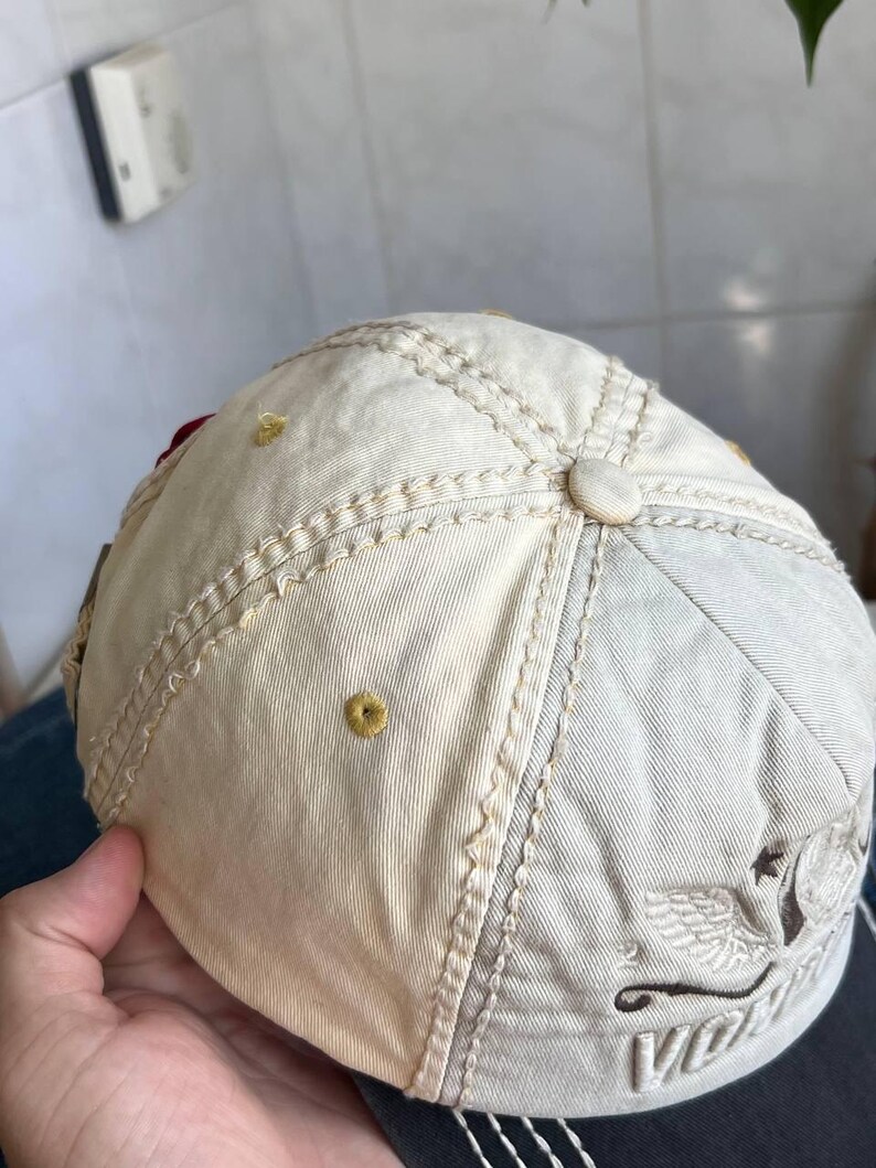 Op de afbeelding: Cr&egrave;mekleurige baseballpet met een gebogen klep. De pet heeft decoratieve stiksels, een knoop aan de bovenkant en een geborduurd ontwerp aan de zijkant. De pet wordt door een hand vastgehouden.