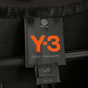 May include: Close-up of a black garment tag with the Adidas logo and the orange "Y-3" logo. The tag also includes the text "YOHJI YAMAMOTO" and size "L/G". The garment is black.