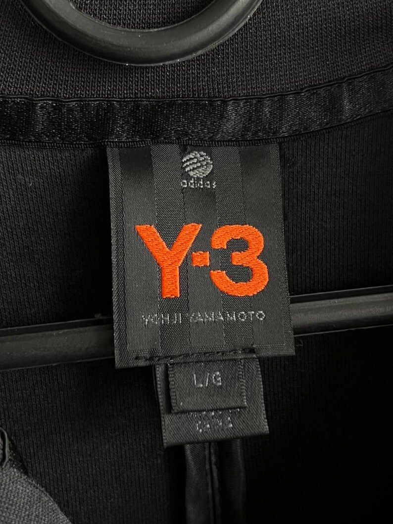 May include: Close-up of a black garment tag with the Adidas logo and the orange "Y-3" logo. The tag also includes the text "YOHJI YAMAMOTO" and size "L/G". The garment is black.
