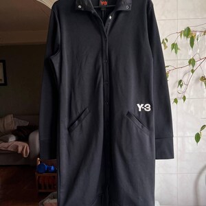 May include: Black long coat with a button-up front, collar, and two front pockets. The coat has the white "Y-3" logo on the front. The coat is hanging on a black hanger.