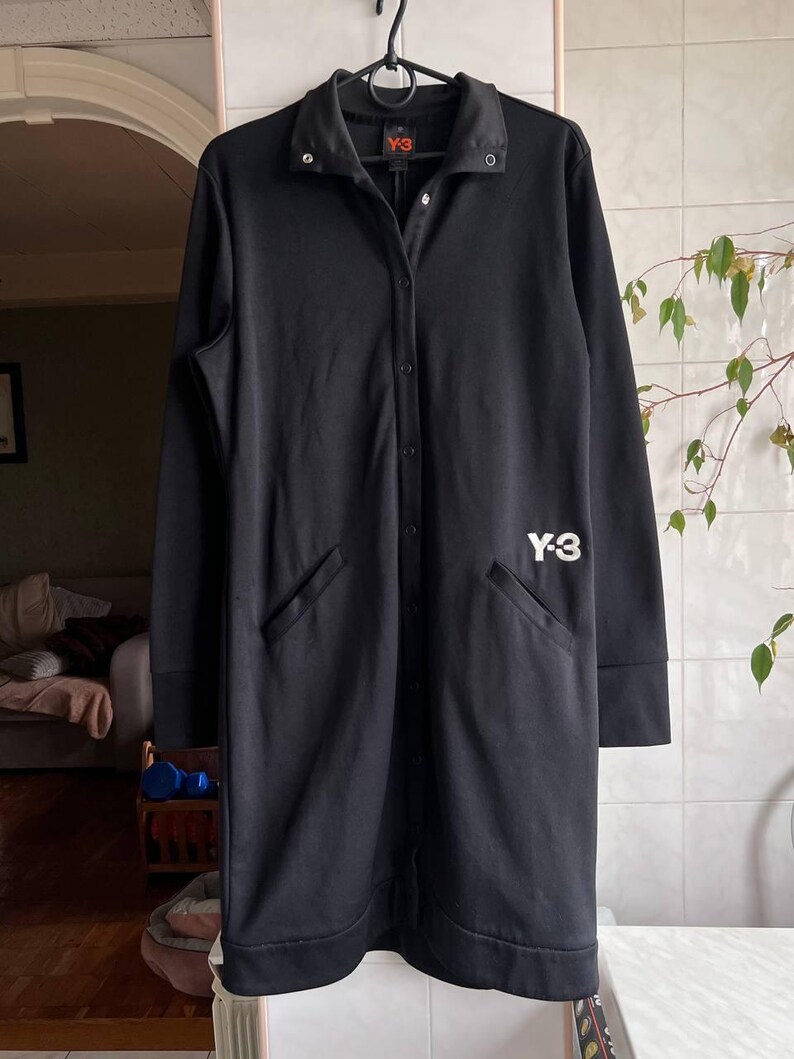 May include: Black long coat with a button-up front, collar, and two front pockets. The coat has the white "Y-3" logo on the front. The coat is hanging on a black hanger.