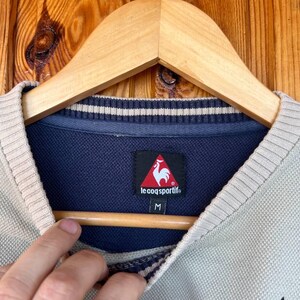 Puede incluir: Primer plano de un su&eacute;ter Le Coq Sportif verde claro y beige. El su&eacute;ter tiene un cuello azul marino con rayas blancas y un logotipo de gallo rojo. La etiqueta de talla dice "M". El su&eacute;ter est&aacute; colgado en una percha de madera.