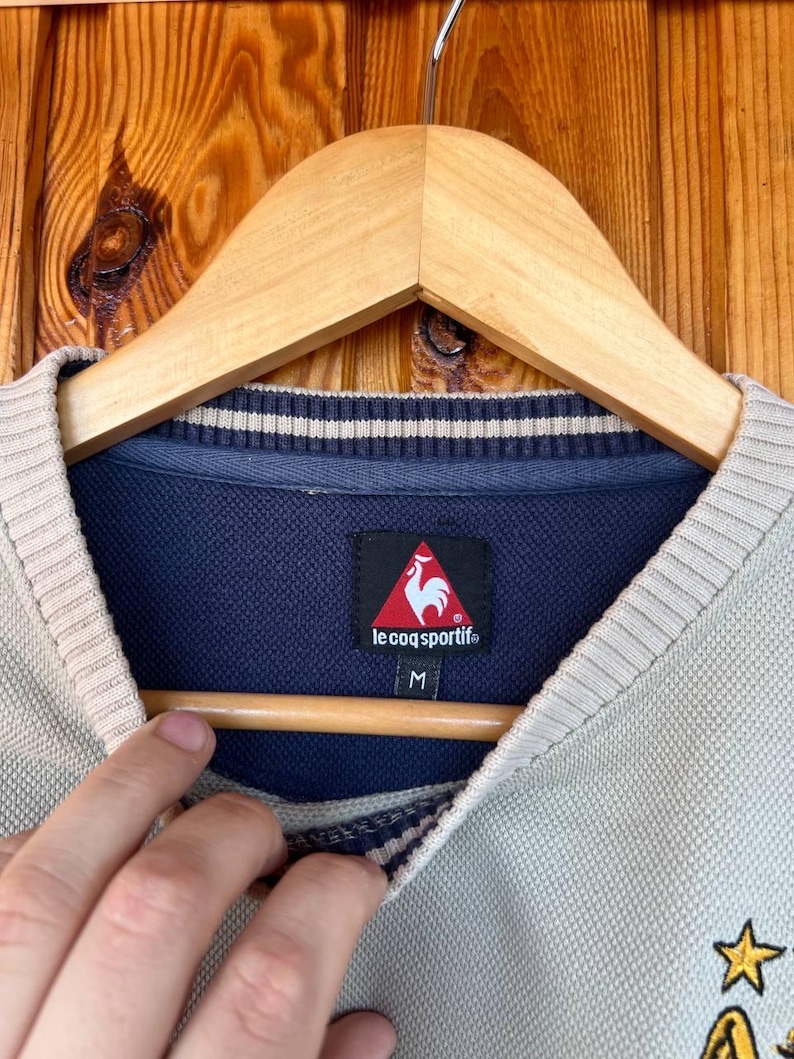Puede incluir: Primer plano de un su&eacute;ter Le Coq Sportif verde claro y beige. El su&eacute;ter tiene un cuello azul marino con rayas blancas y un logotipo de gallo rojo. La etiqueta de talla dice "M". El su&eacute;ter est&aacute; colgado en una percha de madera.