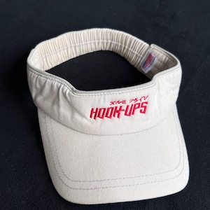 May include: A beige visor with the text "HOOK-UPS" in red, along with Japanese characters. The visor has an elastic band and a curved brim. The visor is made of a soft fabric.