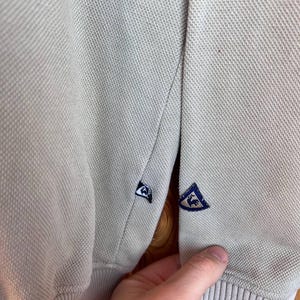 Puede incluir: Primer plano de un polo beige claro. La camisa tiene una tela de punto texturizada y presenta un emblema azul y blanco en la manga. El pu&ntilde;o tiene un detalle de rayas azul marino.