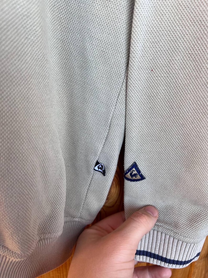 Puede incluir: Primer plano de un polo beige claro. La camisa tiene una tela de punto texturizada y presenta un emblema azul y blanco en la manga. El pu&ntilde;o tiene un detalle de rayas azul marino.