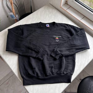 Puede incluir: Una sudadera negra de manga larga con cuello redondo que presenta un pequeño logotipo bordado en el pecho izquierdo. El logotipo incluye el texto "ComputerLand" sobre un icono de manzana multicolor y el texto "Top 30" con "1986" impreso debajo.