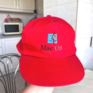 Puede incluir: Una gorra de béisbol de color rojo brillante con el logotipo de Mac OS bordado en la parte delantera. El logo consiste en un cuadrado dividido en un lado azul y otro morado con el contorno de una cara estilizada y el texto Mac OS debajo.