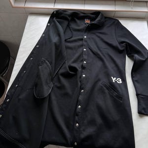 May include: Black button-up jacket with a collar and long sleeves. The jacket has a Y-3 logo on the front and a small logo on the back. The jacket is open, revealing the interior lining and pockets.