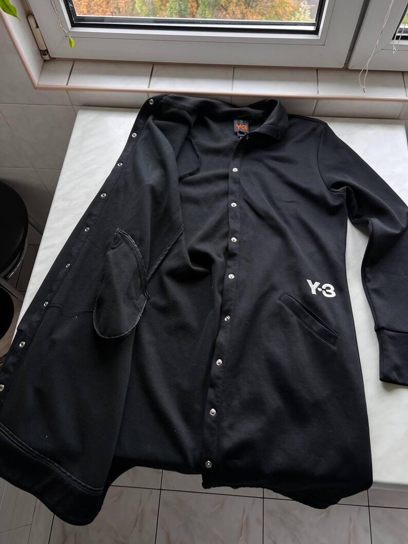 May include: Black button-up jacket with a collar and long sleeves. The jacket has a Y-3 logo on the front and a small logo on the back. The jacket is open, revealing the interior lining and pockets.