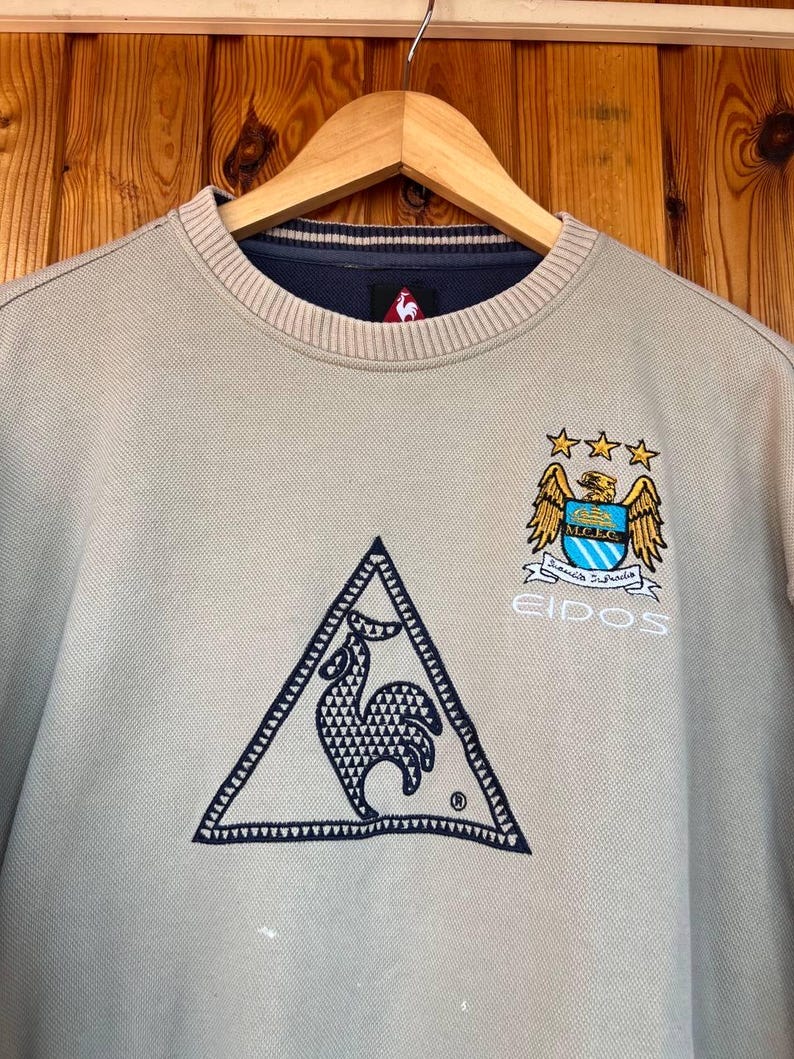 Puede incluir: Sudadera beige de cuello redondo con cuello azul marino. Presenta un logotipo triangular con un gallo, un escudo del equipo con estrellas y la palabra "EIDOS" en blanco. La sudadera est&aacute; colgada en una percha de madera.