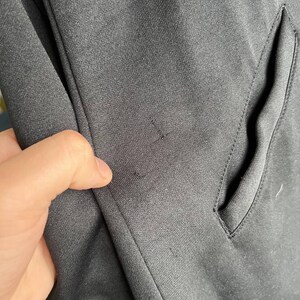May include: Close-up of a black fabric with a pocket and a hand touching the material. The fabric has a subtle texture and appears to be part of a garment, possibly a jacket or coat.