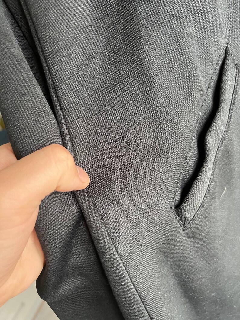 May include: Close-up of a black fabric with a pocket and a hand touching the material. The fabric has a subtle texture and appears to be part of a garment, possibly a jacket or coat.