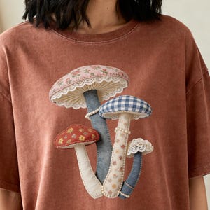 Peut inclure: T-shirt de couleur rouille avec une application de trois champignons décoratifs. Les chapeaux de champignons présentent des motifs floraux, vichy et en dentelle. Le tee-shirt a des manches courtes et une coupe ample, parfait pour un usage quotidien.