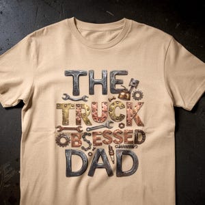Puede incluir: Una camiseta color beige con el texto "THE TRUCK OBSESSED DAD" en un diseño metálico 3D. El texto incorpora llaves inglesas, engranajes y otros elementos mecánicos. La camiseta tiene mangas cortas y cuello redondo.