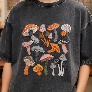 Puede incluir: Una camiseta gris oscuro con un gráfico de setas coloridas. El diseño incluye varias ilustraciones de setas en tonos naranja, marrón, rosa y verde. La camiseta se lleva con un sombrero de paja y pantalones oscuros.
