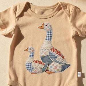 Könnte beinhalten: Ein beiger Baby-Body mit kurzen Ärmeln. Auf der Vorderseite befindet sich ein Applikationsdesign von zwei Gänsen. Die Gänse haben blaue Vichy-, Blumen- und Jeans-Patches. Ein Weidenkorb und eine rosa Decke sind im Hintergrund.
