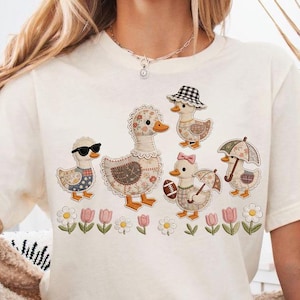 Puede incluir: Camiseta color crema con un diseño caprichoso de seis patos, cada uno adornado con accesorios únicos como gafas de sol, sombreros y lazos. Los patos están colocados sobre un fondo floral. La camiseta se combina con pantalones cortos vaqueros.