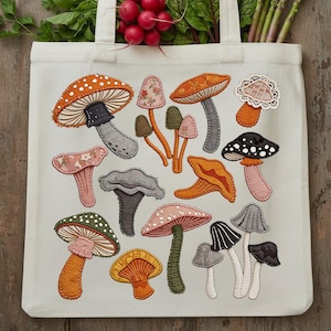 May include: Cream-colored tote bag featuring embroidered mushroom designs in various colors and patterns. The bag is filled with fresh produce, including radishes, spinach, and asparagus. The bag has a natural, rustic aesthetic.