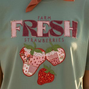 Puede incluir: Camiseta verde salvia con la frase "FARM FRESH STRAWBERRIES" en un diseño de patchwork. Debajo del texto, tres apliques de fresas con detalles florales y de encaje. La camiseta es de manga corta.