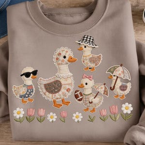 Può includere: Una felpa grigia con applicazioni di anatre con vari accessori, tra cui occhiali da sole e cappelli. Fiori e tulipani ricamati circondano le anatre. La felpa ha un girocollo.