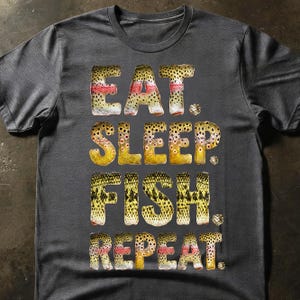 May include: Dark grey t-shirt featuring the text "EAT. SLEEP. FISH. REPEAT." in a font designed to look like fish scales. The text is in shades of yellow, brown, and red. A casual tee for fishing lovers.