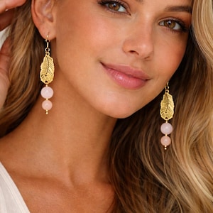 Peut inclure: Boucles d'oreilles pendantes en forme de plume dorée avec deux perles roses. Les boucles d'oreilles ont un design délicat. Elles sont portées par une personne aux cheveux blonds et un haut blanc.