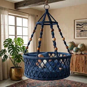 May include: A navy blue macrame baby swing with wooden beads, suspended from the ceiling. The swing has a circular design and a soft blue cushion. A baby is inside the swing. The room has a bohemian style with a large potted plant and a wooden cabinet.