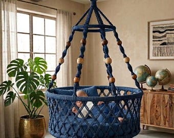 May include: A navy blue macrame hanging cradle featuring a circular base with a diamond-patterned net, suspended by thick knotted ropes adorned with natural wooden beads. A small infant rests inside on a matching navy blue cushioned mattress.