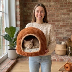 May include: A dome-shaped, rust-colored crocheted cat bed with a wide arched opening. Inside, a light-colored cushion features a pattern of small gray paw prints. A ginger cat is resting comfortably within the interior of the soft, textured structure.