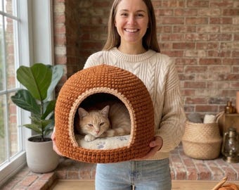 May include: A dome-shaped, rust-coloured crocheted cat bed with a wide arched opening. Inside, a light-coloured cushion features a pattern of small grey paw prints. A ginger cat is resting comfortably within the interior of the soft, textured structure.