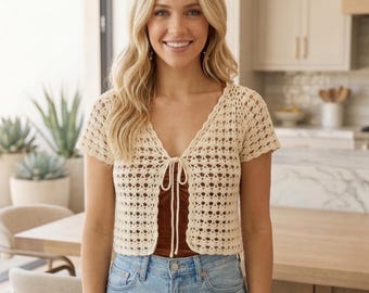 May include: A cream-coloured, short-sleeved crochet cardigan with a V-neck and tie closure. The openwork design reveals a brown top underneath. The cardigan is paired with blue jeans, creating a casual, everyday look.