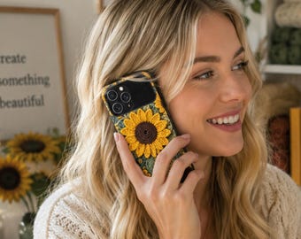 May include: A smartphone case featuring a textured, crocheted sunflower design with a dark brown centre and bright yellow petals. The case has a black camera cutout area and a green, leafy patterned background surrounding the central sunflower motif.