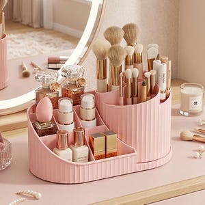 Puede incluir: Un organizador de maquillaje rosa y acanalado lleno de brochas, perfumes y cosméticos. El organizador se encuentra sobre una superficie rosa, con un espejo en el fondo. También se ven una vela y una scrunchie.