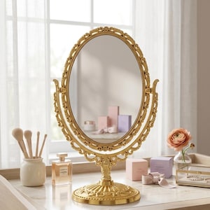 Puede incluir: Un espejo de tocador ovalado dorado con detalles ornamentados. El espejo está sobre un soporte decorativo. Pinceles de maquillaje, perfume y otros productos de belleza están colocados en la mesa de tocador. Una rosa en un jarrón de cristal añade color.