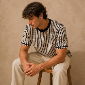 May include: A short-sleeved, striped shirt with a navy blue collar and cuffs. The shirt features vertical black stripes on a cream-colored background. The person is wearing cream-colored pants and is seated on a wooden stool. The overall style is casual.