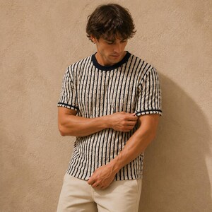 May include: A short-sleeved, cream-colored shirt with vertical black stripes. The shirt has a navy blue crew neck and sleeve trim. The person in the image is wearing khaki-colored pants.