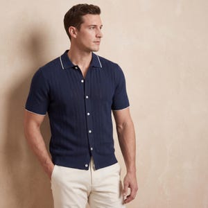 May include: Navy blue short-sleeve shirt with white trim on the collar and sleeves. The shirt has a ribbed texture and is buttoned down the front. The person is wearing light-colored pants.