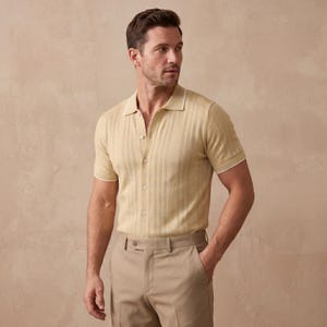 May include: A light tan short-sleeve polo shirt with a textured vertical stripe design and white trim on the collar and sleeves. The shirt is buttoned up and paired with matching tan trousers. The outfit is a classic, casual style.