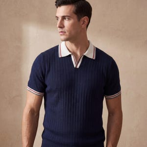 Puede incluir: Polo de punto acanalado azul marino con cuello blanco y ribete rojo y gris en el cuello y las mangas. La camisa tiene un dise&ntilde;o de cuello en V.