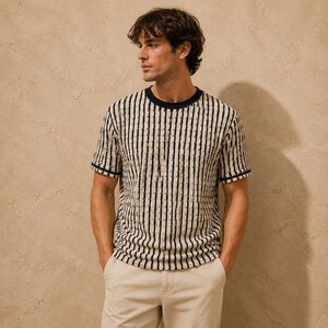May include: A short-sleeved, striped t-shirt with a navy blue neckline and sleeve trim. The shirt features vertical black stripes against a cream-colored background. The model is wearing khaki-colored pants.