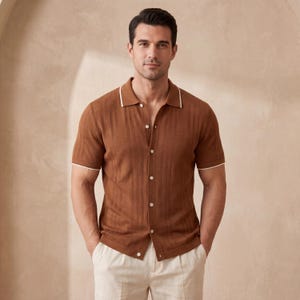 Puede incluir: Una camisa marrón de manga corta con un diseño texturizado y ribete blanco en el cuello y las mangas. La camisa está abotonada y se combina con pantalones de color claro. El estilo general es casual y elegante.