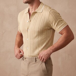 May include: A light tan polo shirt with a textured vertical stripe design. The shirt has a white collar and sleeve trim, and is buttoned up. The model is wearing khaki pants. The shirt is made of a lightweight material.