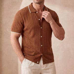 May include: A brown short-sleeved polo shirt with a textured pattern and white trim on the collar and sleeves. The shirt has a button-down front and is paired with light-colored pants. The shirt is made of a knit fabric.