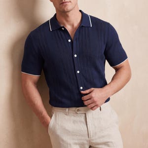 May include: Navy blue short-sleeve polo shirt with white trim on the collar and sleeves. The shirt has a textured design and is buttoned up. The model is wearing beige pants.
