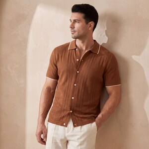 May include: A brown short-sleeve polo shirt with white trim on the collar and sleeves. The shirt has a button-down front and a textured design. The model is wearing off-white pants.