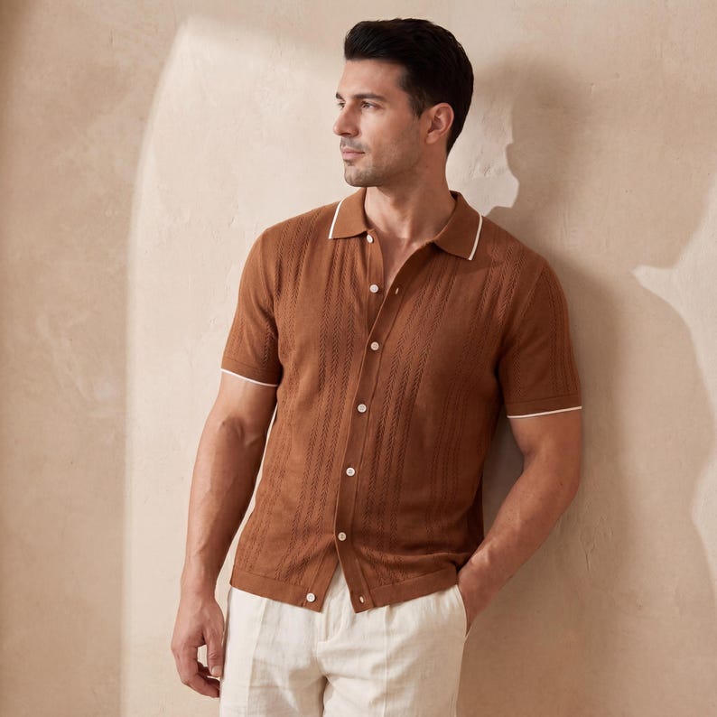 May include: A brown short-sleeve polo shirt with white trim on the collar and sleeves. The shirt has a button-down front and a textured design. The model is wearing off-white pants.