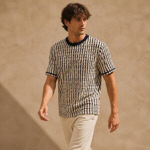 May include: A short-sleeved, striped t-shirt in cream and black. The shirt has a crew neck and the edges of the sleeves are trimmed with a navy blue stripe. The model is wearing cream-colored pants.